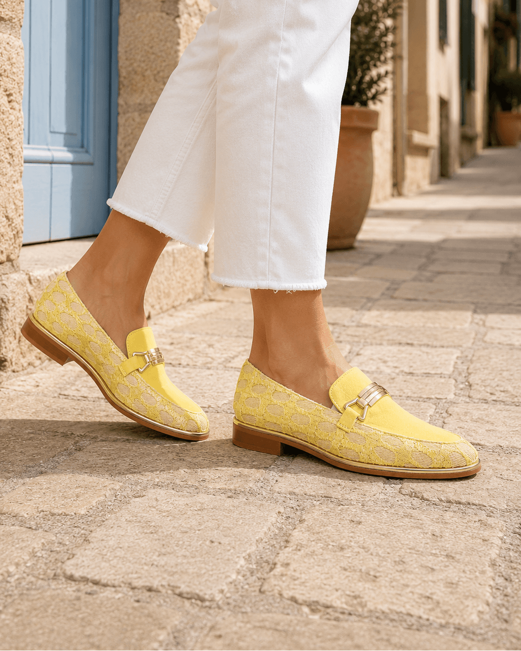A person wearing bright yellow patterned loafers with gold-tone buckle details and white cropped jeans, standing on a stone street in a sunny outdoor setting.